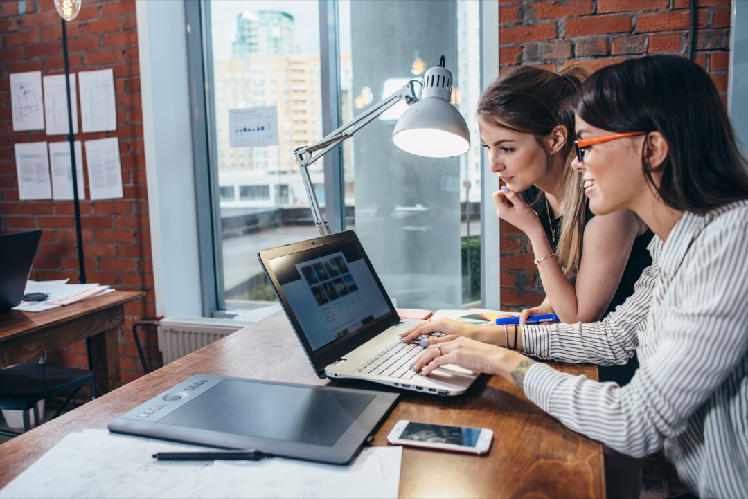 Two women working on new website design choosing pictures using the laptop surfing the internet.